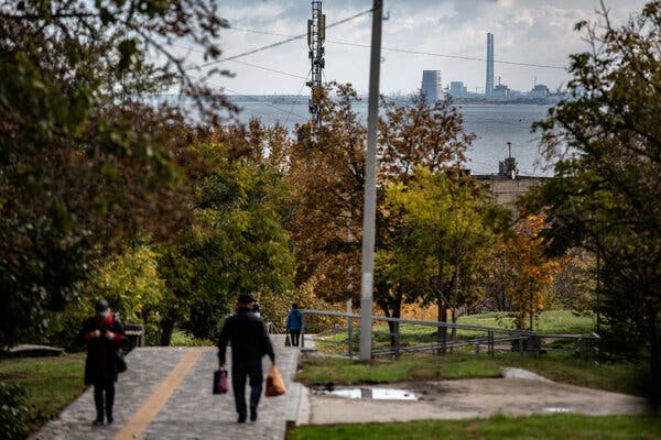 На задньому плані атомна електростанція навпроти водойми. На передньому плані люди йдуть міським парком.